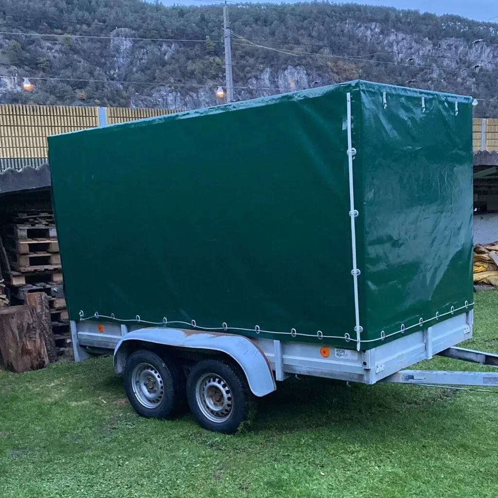 Anhänger mit grüner Plane auf Rasen vor bewaldeter Kulisse.