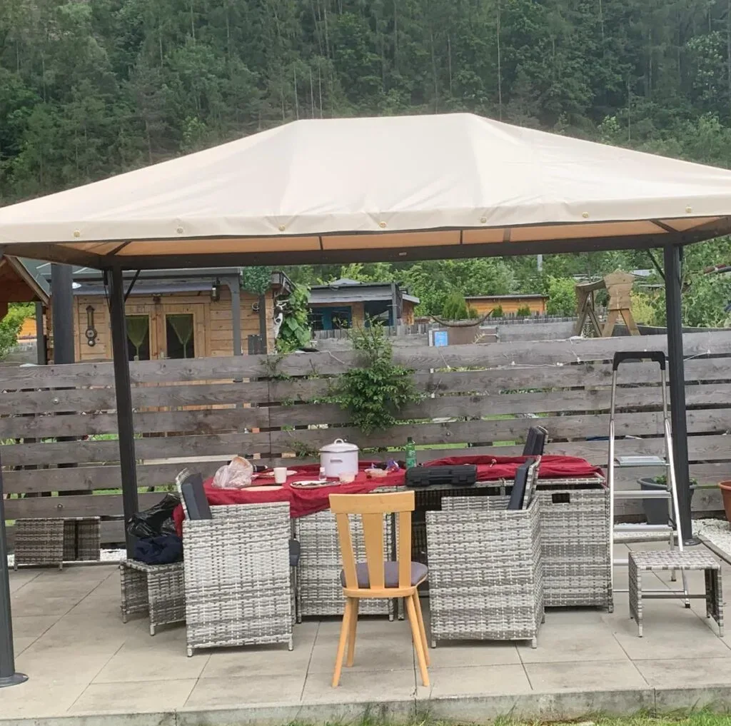 Pavillon mit beigem Dach über Esstisch und Stühlen, umliegende Pflanzen und Holzzaun.
